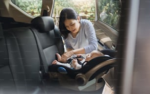 Asian young mother putting her baby son into car seat and fasten seat belts in the car for safety in transportation.
