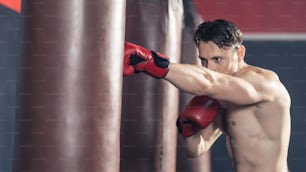 Caucasian bodybuilder man shirtless athlete wear boxing gloves, doing workout exercise by hitting a punching bag or Boxing Sandbag. Young sportsman boxer maintain muscle for health in gym or fitness.