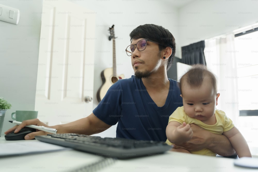 Padre asiático trabajando desde casa y cuidando a su pequeño hijo. Hombre diseñador usando computadora durante la cuarentena.