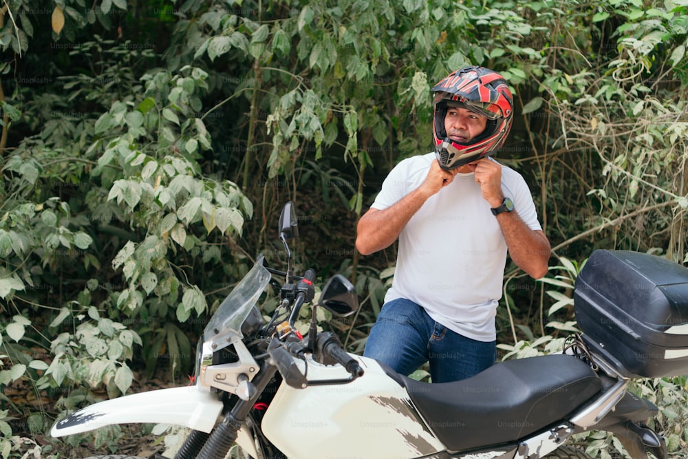 Young man in white shirt and jeans strapping on a helmet on a motorcycle in the open air.