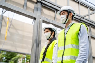 Asiatische Arbeiter tragen Schutzmasken auf einer Baustelle aufgrund der Covid-19- oder Coronavirus-Epidemie, männliche und weibliche Ingenieure, die sich das Gebäude ansehen, Bauarbeiten durchführen, über das Projekt sprechen.