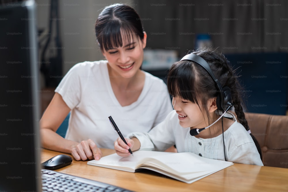 コロナウイルスのパンデミックによるデジタルリモートインターネット会議によって学校の先生からオンラインクラスを学んでいるホームスクーリングアジアの若い女の子。子供がパソコンを見てメモを書き、お母さんと一緒に座っている。