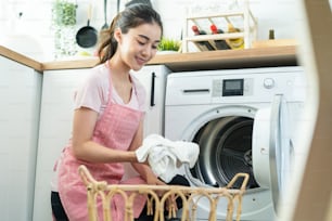 Asian beautiful woman put dirty clothes to washing machine in house. Attractive girl wear apron sit on floor, feel happy to loads laundry in washer appliance at home. Domestic-House keeping concept.