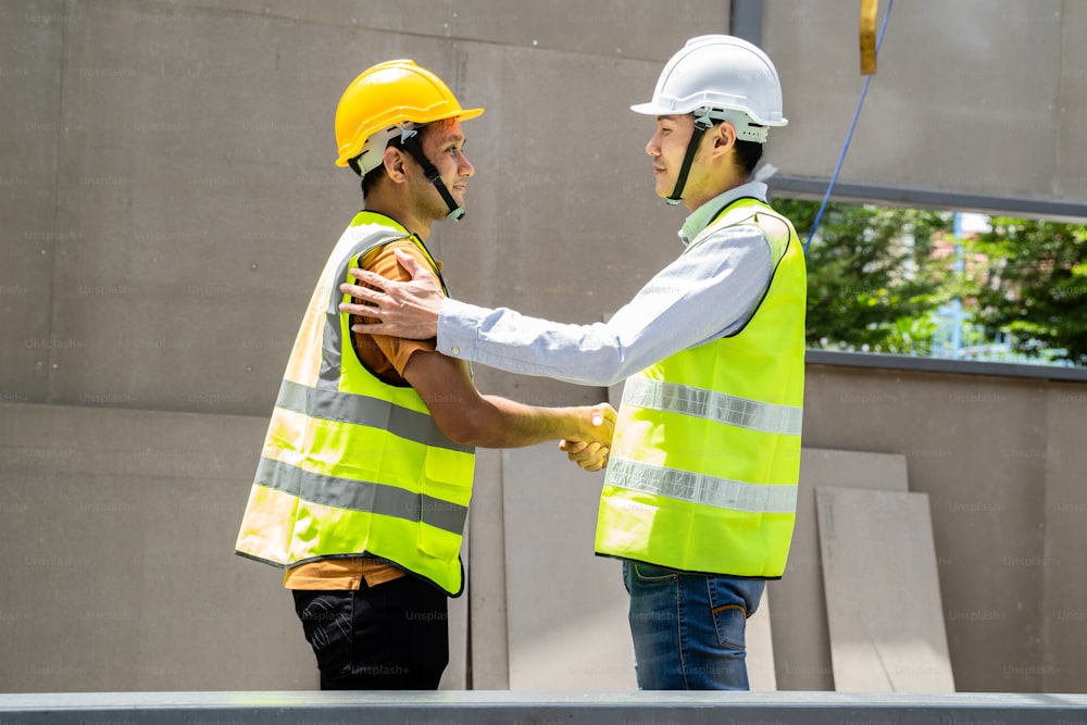 建築現場で握手を交わすアジアの構造エンジニアと労働者。建設現場を歩くときに安全ヘルメットをかぶっているすべての人。商取引、M&Aのコンセプト