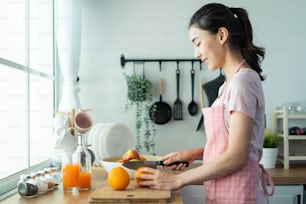 Asian attractive woman make drinking orange juice in kitchen at home. Smiling young beautiful girl wear apron feel happy enjoy cook healthy foods and drink to lose weight and diet for health in house.
