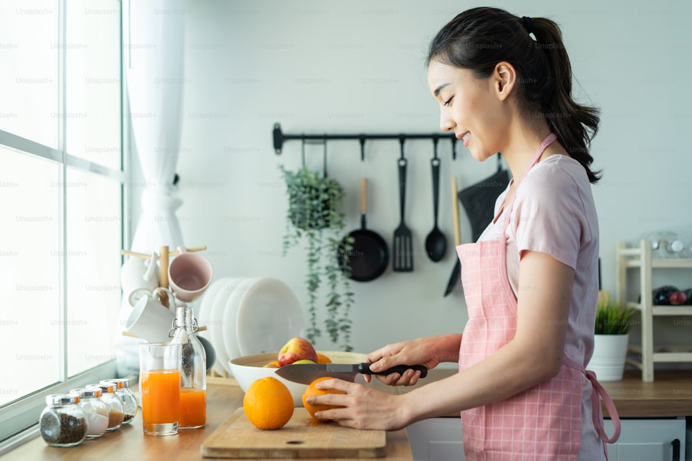 La donna attraente asiatica fa bere succo d'arancia in cucina a casa. La giovane ragazza sorridente e bella indossa il grembiule si sente felice, si diverte a cucinare cibi sani e a bere per perdere peso e la dieta per la salute in casa.