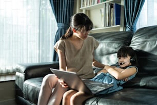 Asian mom motivate young girl student daughter to learn online at home. Unhappy little cute kid give up from virtual online study class from school teacher due to Covid-19 and learn on sofa in house.