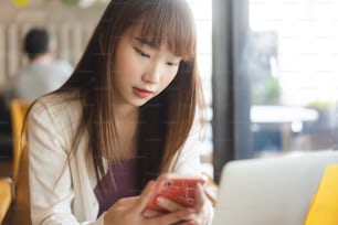 大学の人々、教育、ライフスタイル、仕事、昼間のコンセプトに関する研究。屋内カフェでのオンライン申請に携帯電話を使用する若い大人のアジアの女子学生。