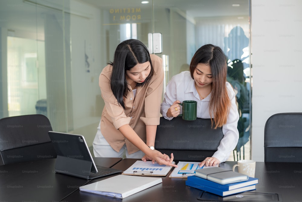 아름다운 두 젊은 아시아 사업가 브레인 스토밍은 그래프를 사용하여 협업을 분석하고 사무실에서 커피를 마신다.