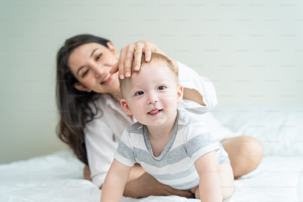 Portrait of Caucasian happy family smiling, look at camera in bedroom. Young attractive woman parents, mother in pajamas laying on bed with little baby boy child enjoy morning wake up activity at home