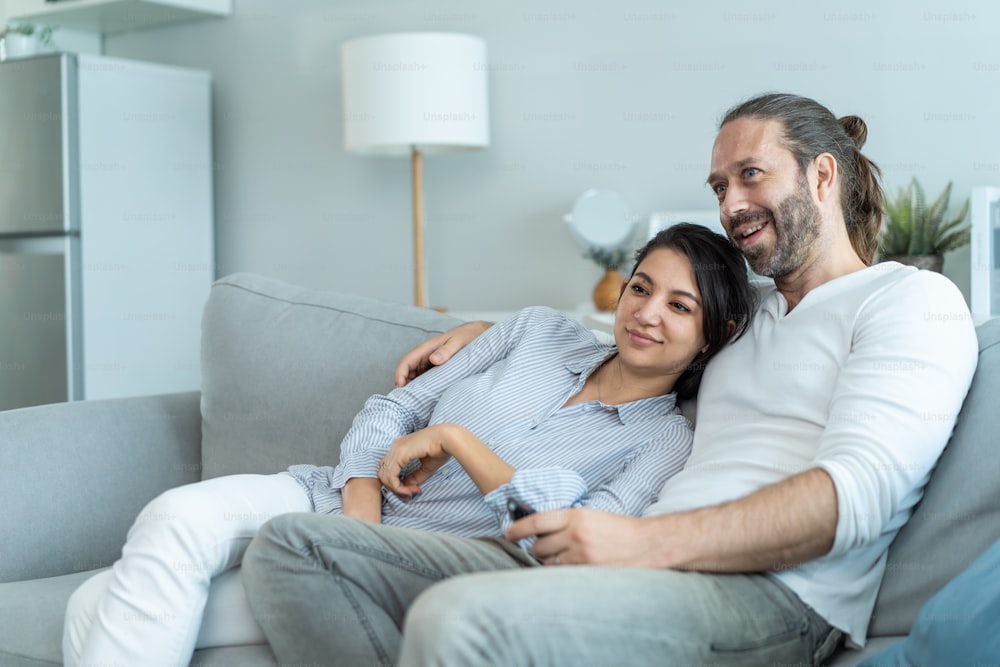 Caucasian loving couple watch movie together in living room at home. Beautiful happy new marriage man and woman sit on sofa use remote control TV show and have fun laughing look to television in house