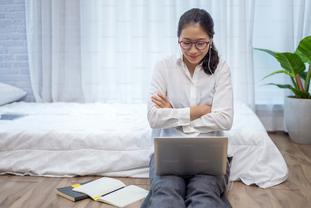 Une jeune femme asiatique porte des écouteurs pour parler et faire des vidéoconférences sur un ordinateur portable dans la chambre à coucher à la maison.