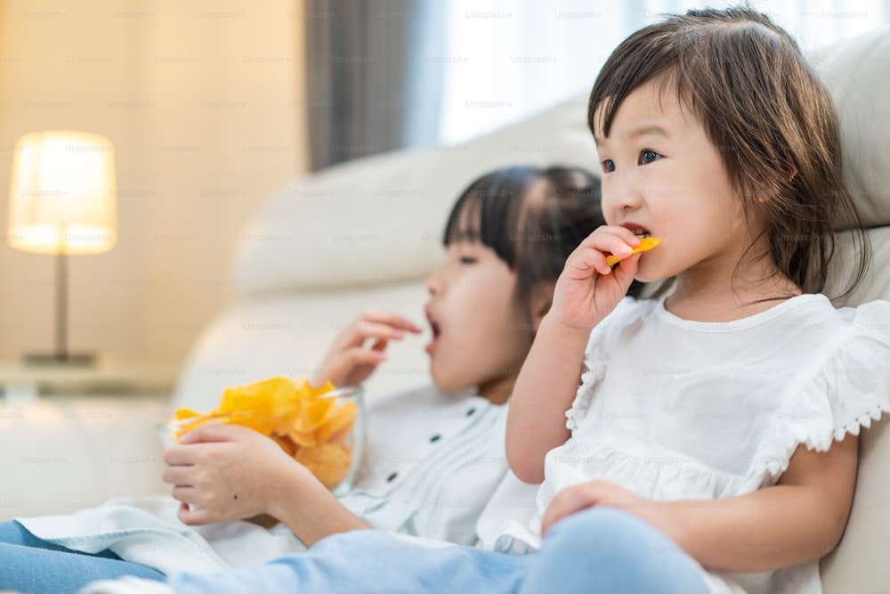 Asian Hungry little girl sibling sisters Puts snack in mouth with hand. Two sweet preschool kid resting on sofa at home enjoy eat fast food, potato chips. Tasty and unhealthy food for children concept