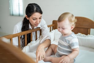 Caucasian mother play with cute baby boy child on infant bed at home. Happy family, attractive young woman mom read book to toddler son with happiness enjoy after wake up activity in bedroom in house.