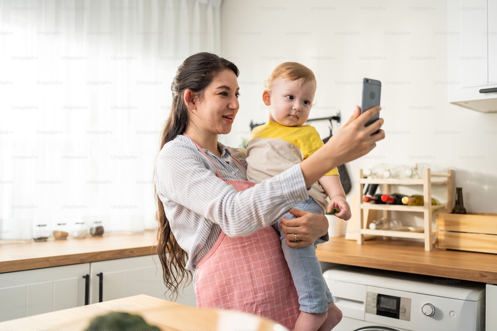 Caucasian mother holding little baby and video call online with family. Attractive beautiful mom using mobile cellphone and chat with husband while carrying young toddler kid in living room in house.