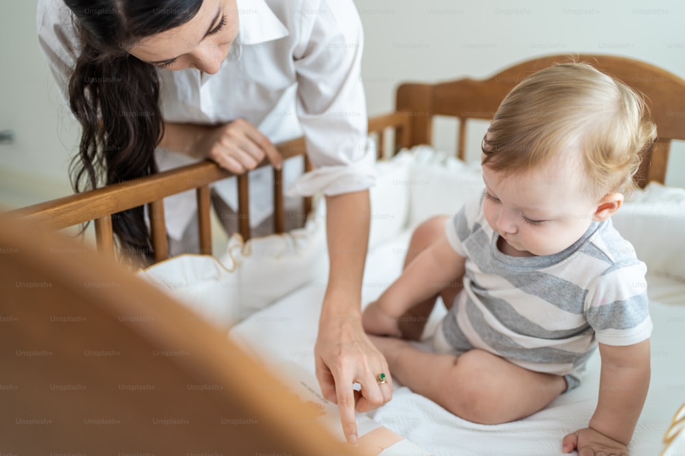 Caucasian mother play with cute baby boy child on infant bed at home. Happy family, attractive young woman mom talking to toddler son with happiness enjoy after wake up activity in bedroom in house.