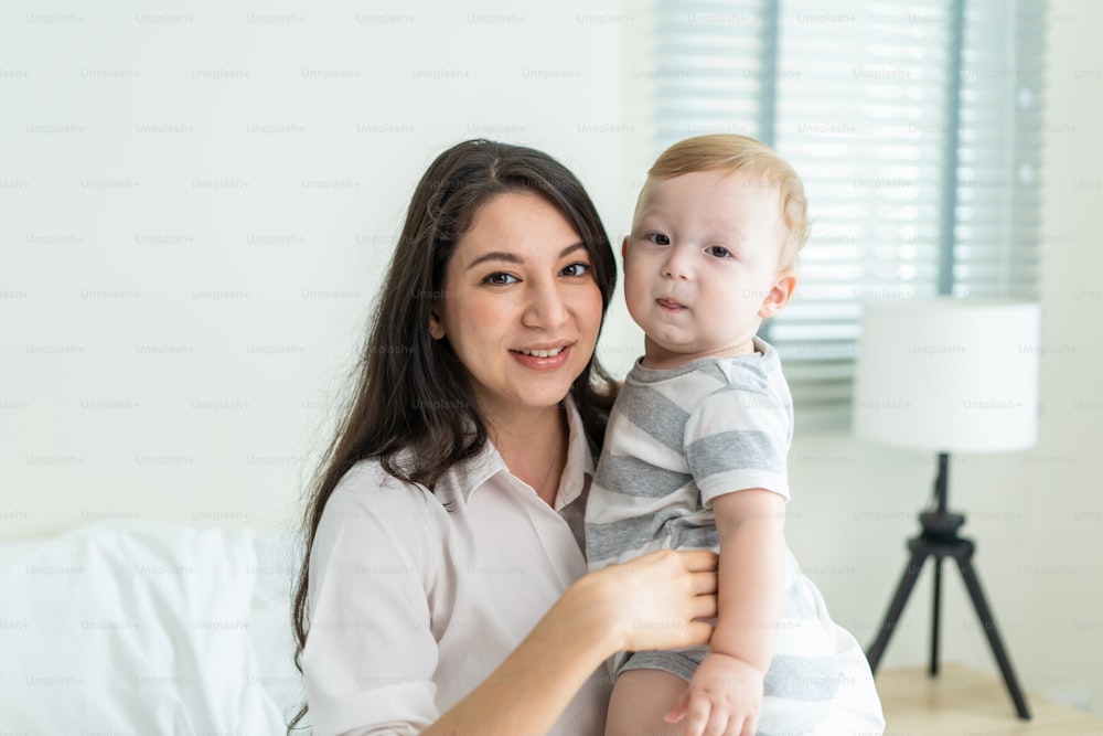 Portrait of Caucasian happy family smiling, look at camera in bedroom. Young attractive woman parents, mother in pajamas sit on bed with little baby boy child enjoy morning wake up activity in house.
