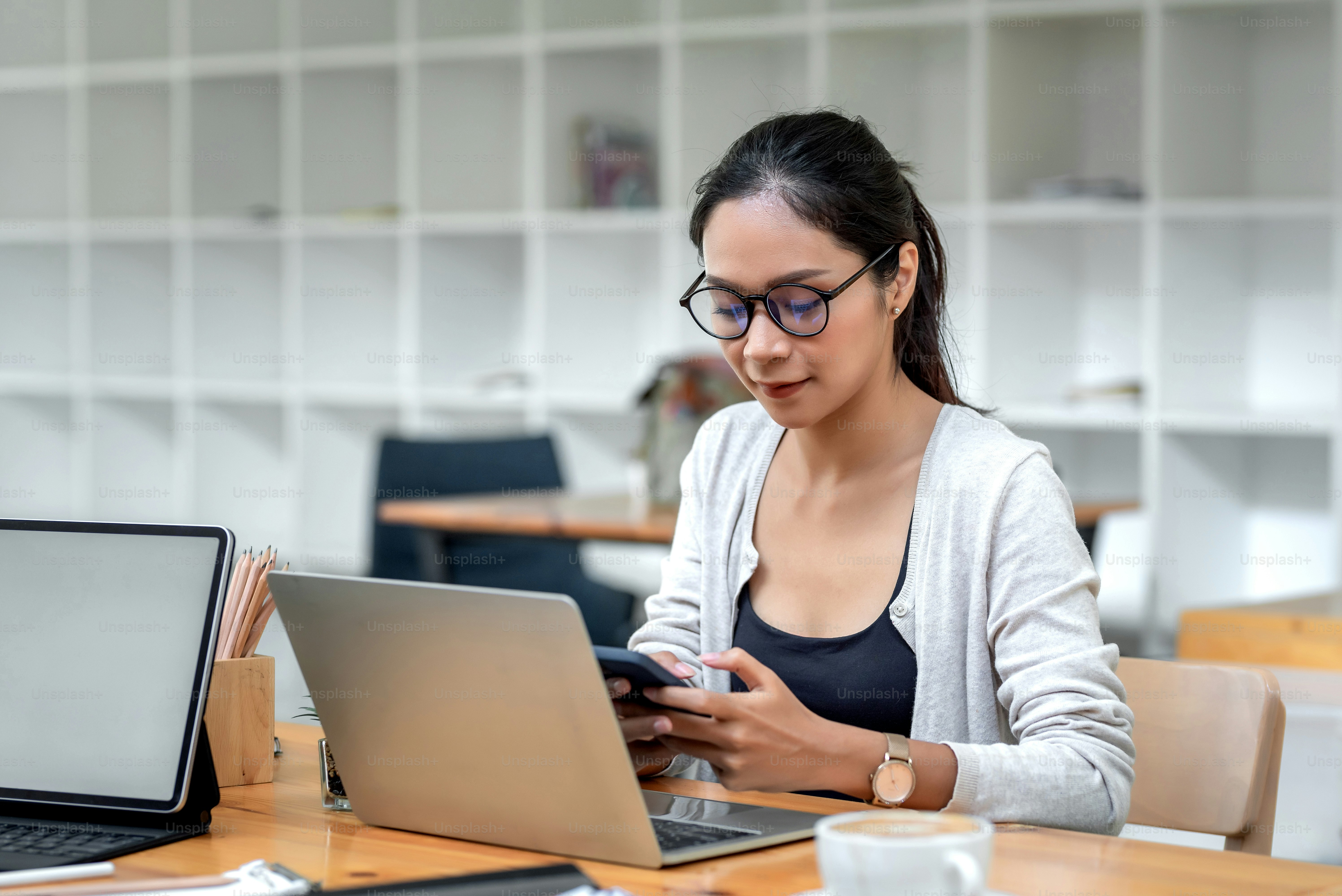 Pengusaha muda Asia berkacamata duduk di kantor sambil memegang smartphone di meja.