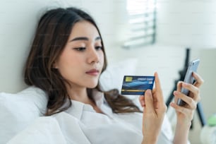 Asian attractive woman enjoy shopping online on bed at home in morning. Casual female lying in bedroom, holding credit card to make e bank online payment after use phone purchase in web store in house