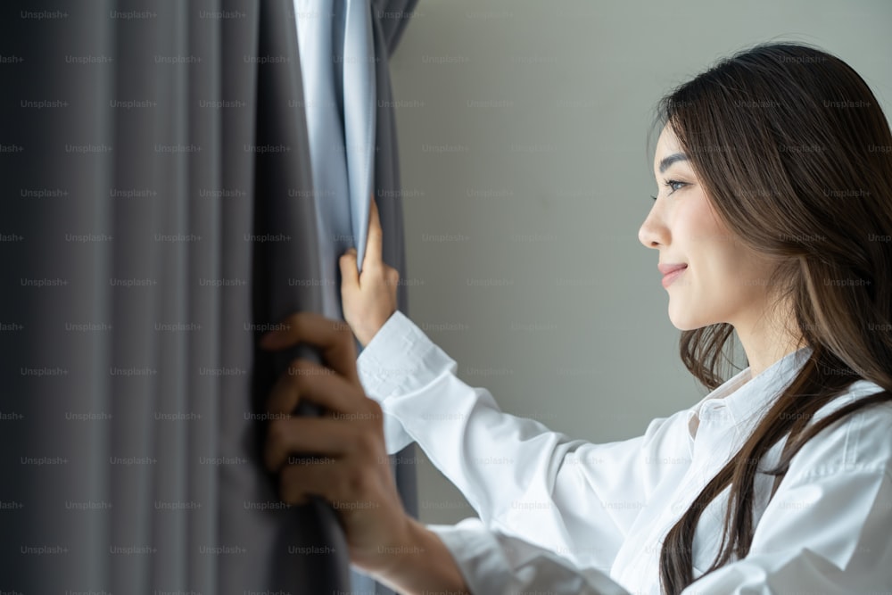 Asian beautiful woman approaches a window and open curtains in morning. Young attractive girl in white pajamas feel fresh and happy to look outside and sunrise after wake up early in bedroom at home.