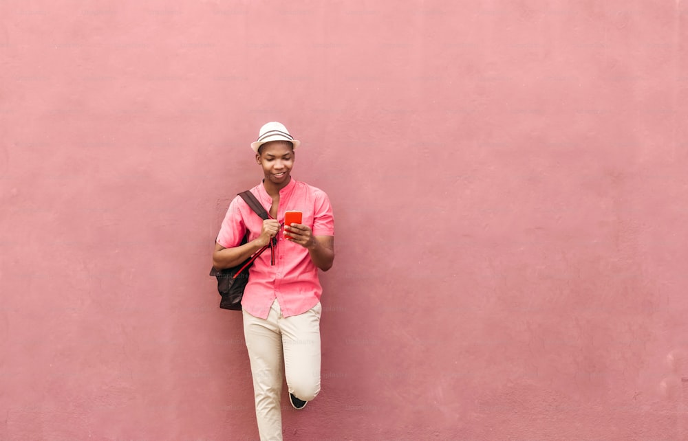 Uomo latino-americano con zaino e cappello in piedi su una parete colorata che guarda un cellulare per smartphone.