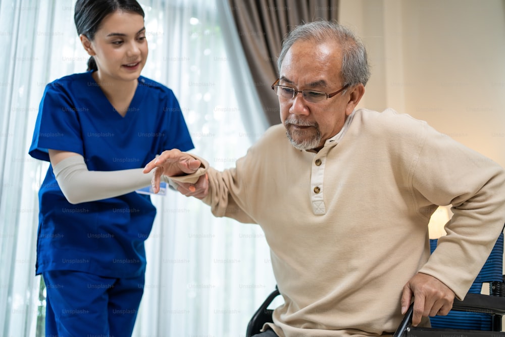 Asian Caregiver Mädchen Arzt Unterstützung älteren behinderten Mann gehen im Haus. Attraktive Krankenschwester, die sich um reife ältere ältere männliche Patienten kümmert, die im Rollstuhl sitzen, häusliche Pflege und Therapie.
