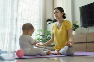 Asian Young mother doing yoga in meditation with baby on yoga mat in the Living room at home.