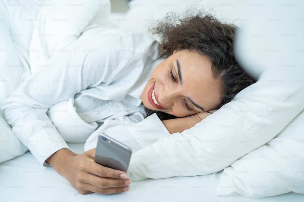 Attractive Latino girl use mobile phone chat on bed at home in morning. Happy casual beautiful female lying down in bedroom enjoy holiday weekend, use smartphone communicate with technology in house.
