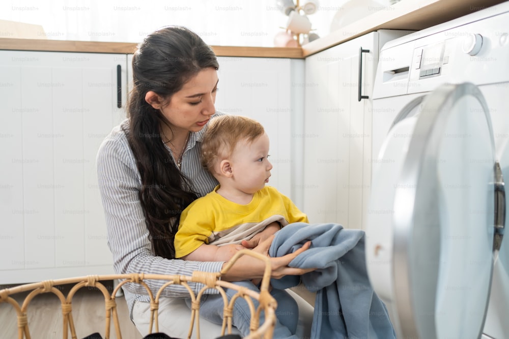 Caucasian busy mother doing housework with baby boy toddler in kitchen. Beautiful young mom putting clothes to washing machine while her little child son playing around in house. Family-housekeeping.