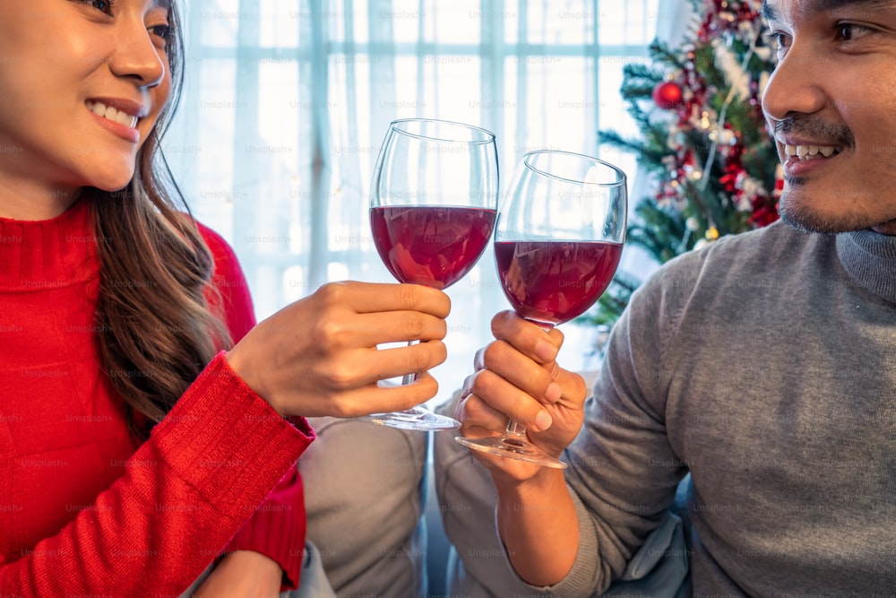 Asian young happy couple celebrate christmas party together in house. Attractive beautiful man and woman drinking red wine alcohol and toasting glass on sofa, feel happy and excited for Xmas at home.