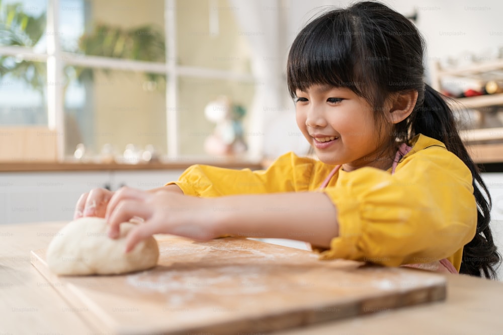 Nahaufnahme asiatisches glückliches junges Kind, das hausgemachte Bäckerei in der Küche macht. Entzückendes kleines Kind sitzt auf dem Tisch und fühlt sich glücklich und genießt es, Essen zu kochen oder zu backen knetet Hefeteig mit den Händen zu Hause