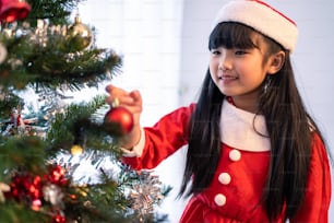 Asian young happy girl kid wear Santa hat and decorate Christmas tree. Young little adorable child wearing red cloth feel excited to celebrate holiday X-mas thanksgiving party in living room in house.