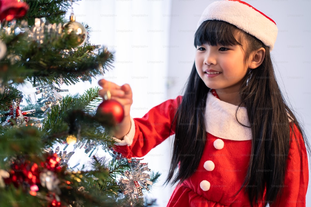 Asian young happy girl kid wear Santa hat and decorate Christmas tree. Young little adorable child wearing red cloth feel excited to celebrate holiday X-mas thanksgiving party in living room in house.