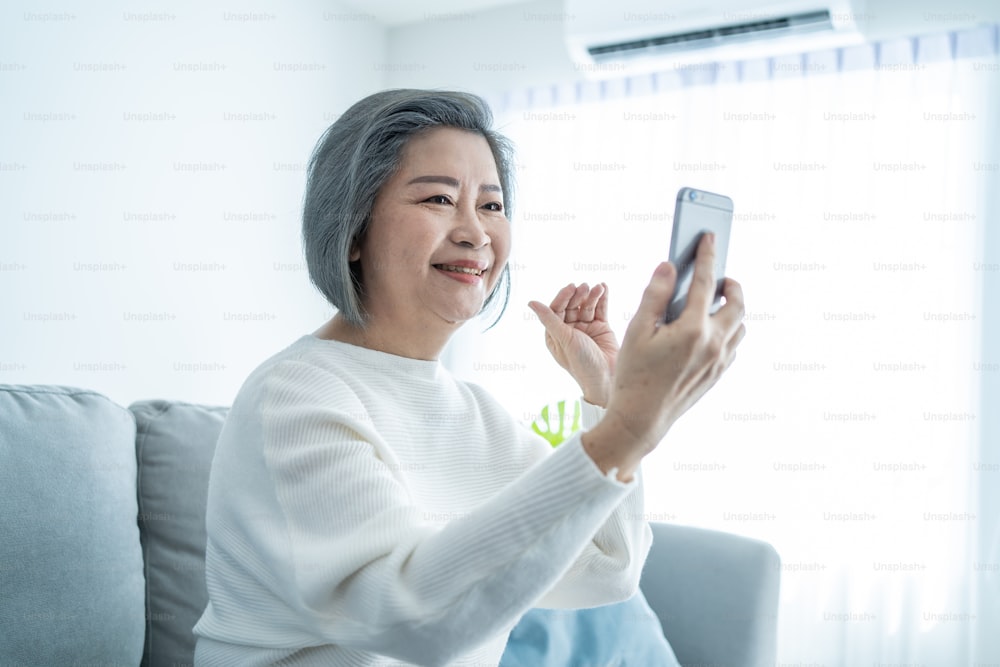 Asian senior elderly woman smile and video call in living room at home. Strong elderly older grandmother feeling happy using mobile cell phone communicate with family enjoy retirement life in house.