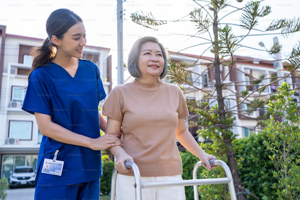 Asian caregiver nurse support old disabled woman to walking outdoors. Beautiful girl doctor help and take care of senior elderly mature handicap patient doing physical therapy in park at nursing home.