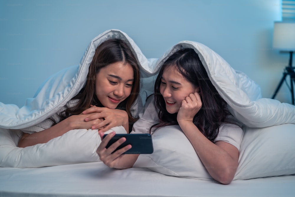 Asian attractive lesbian couple use mobile phone watch movie on bed. Beautiful sibling in pajamas lying down with blanket in bedroom together, use smartphone look at video online at night in house.