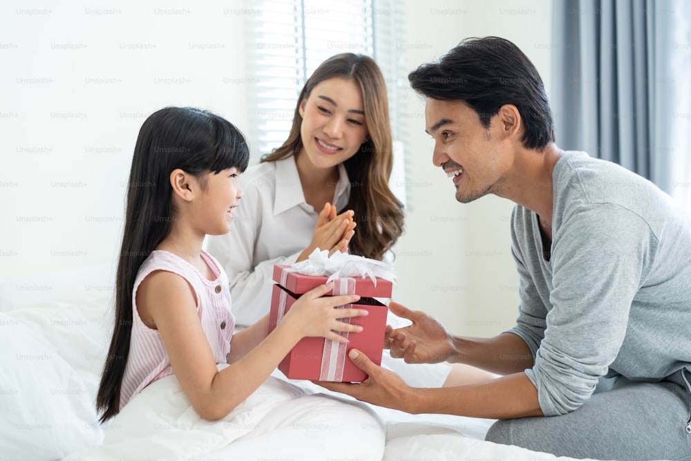 Asian lovely young little daughter open present gift box from parents. Happy family, kid feel happy and excited for surprise from mother and father ready to celebrate Birthday party together at home.