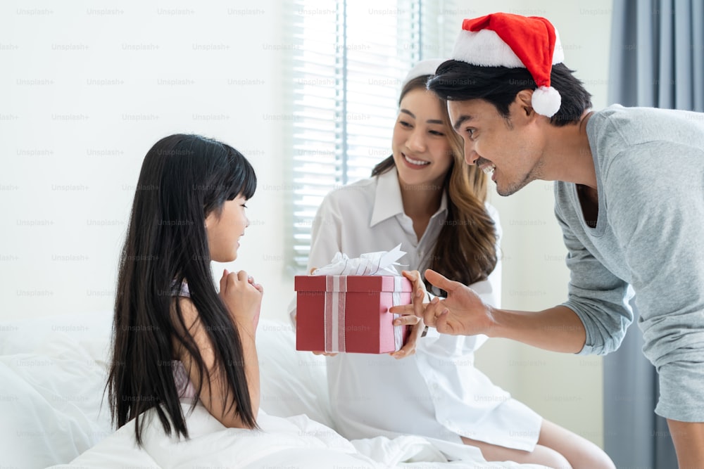 Asian lovely young little daughter open present gift box from parents. Happy family, little kid feel happy and excited for surprise from mother and father ready to celebrate Christmas together at home