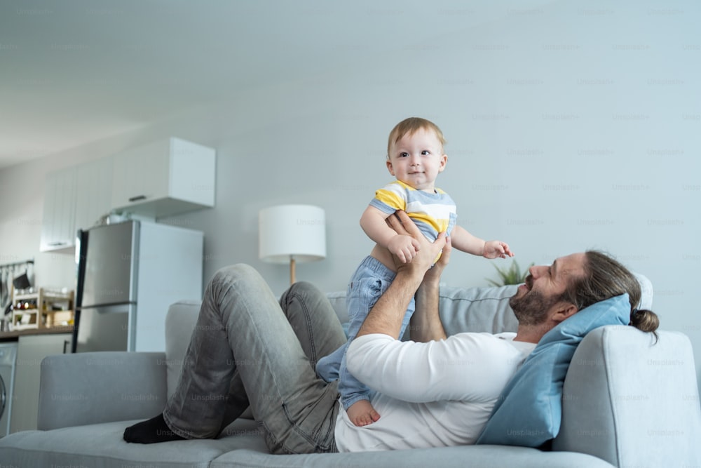 Caucasian loving dad play with baby boy child on sofa in living room. Happy family, attractive handsome caring young father lifting toddler son fly up enjoy doing airplane game in morning in house.