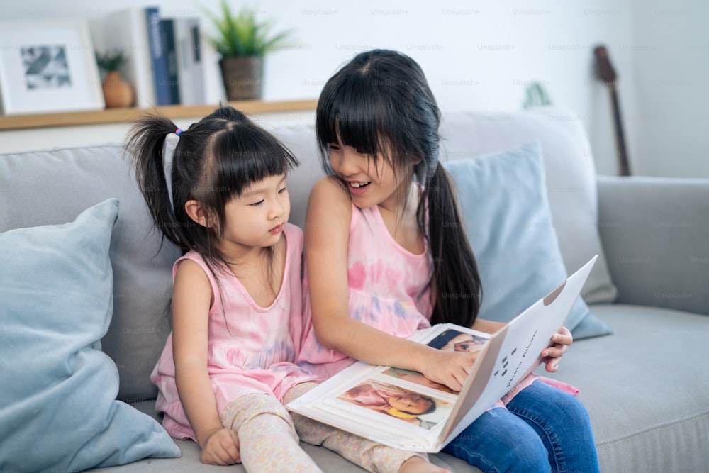 Asian big sister showing old album recalls the past to younger sibling. Young adorable children girl sitting on sofa, flips pages of photo book and enjoy remembering relationship memory in photographs