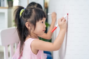 Asian young sibling kid girl enjoy paint on white wall in living room. Little adorable children having fun drawing and coloring art picture with hapiness enjoy creativity activity on holiday at home.