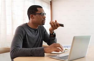 Freelance masculin afro-américain sérieux parlant smartphone à l’aide de la reconnaissance vocale et travaillant sur un ordinateur portable sur le lieu de travail.