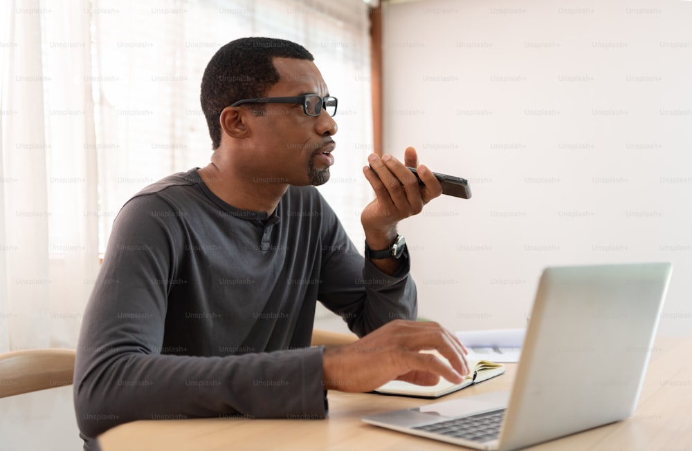Sério afro-americano Freelancer falando smartphone usando reconhecimento de voz e trabalhando em laptop no local de trabalho.