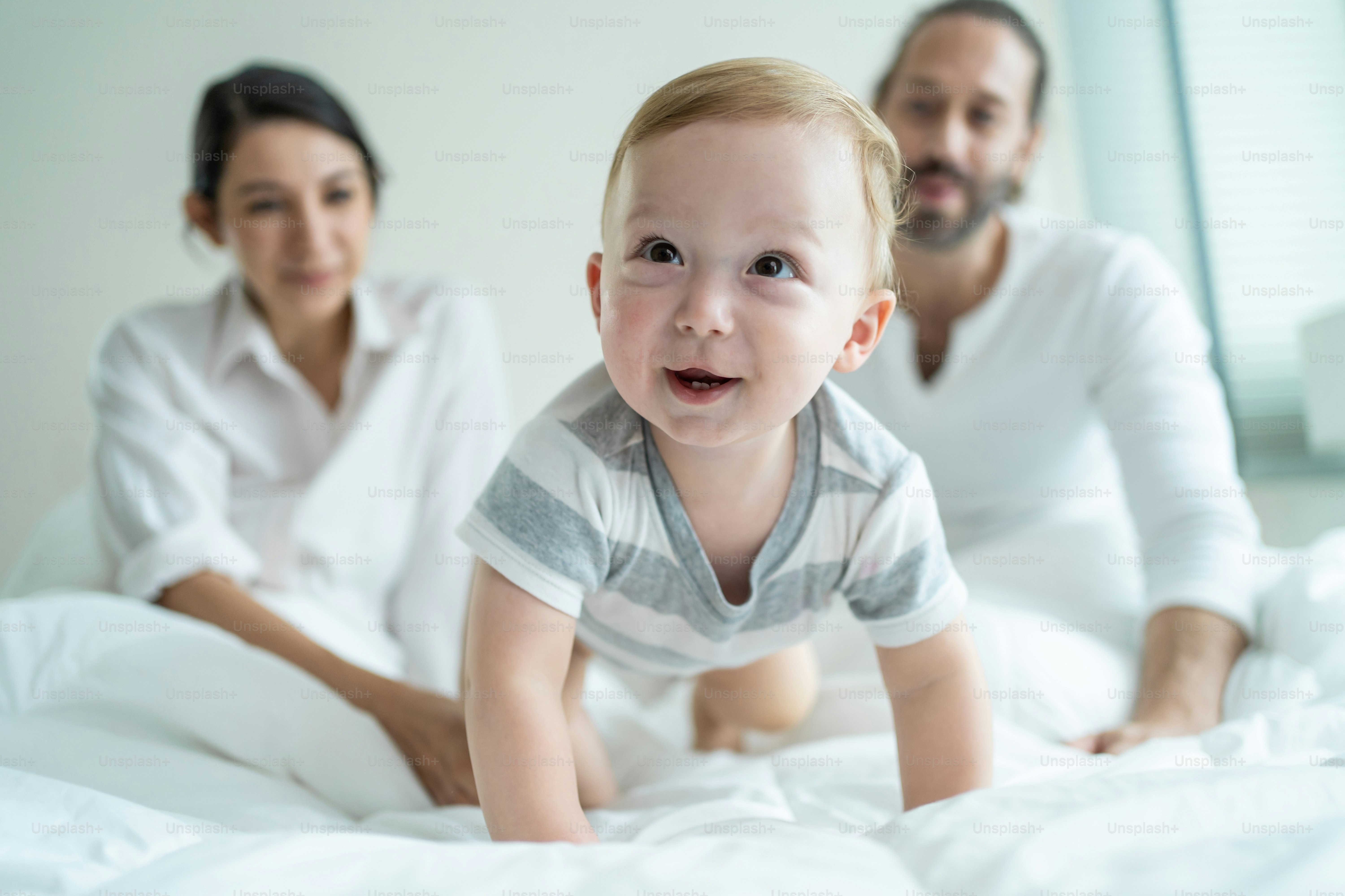 Caucasian parents playing with cute baby boy child on bed in bedroom. Happy family, attractive beautiful caring young mother and father feel happy look at toddler kid son crawling in morning in house.