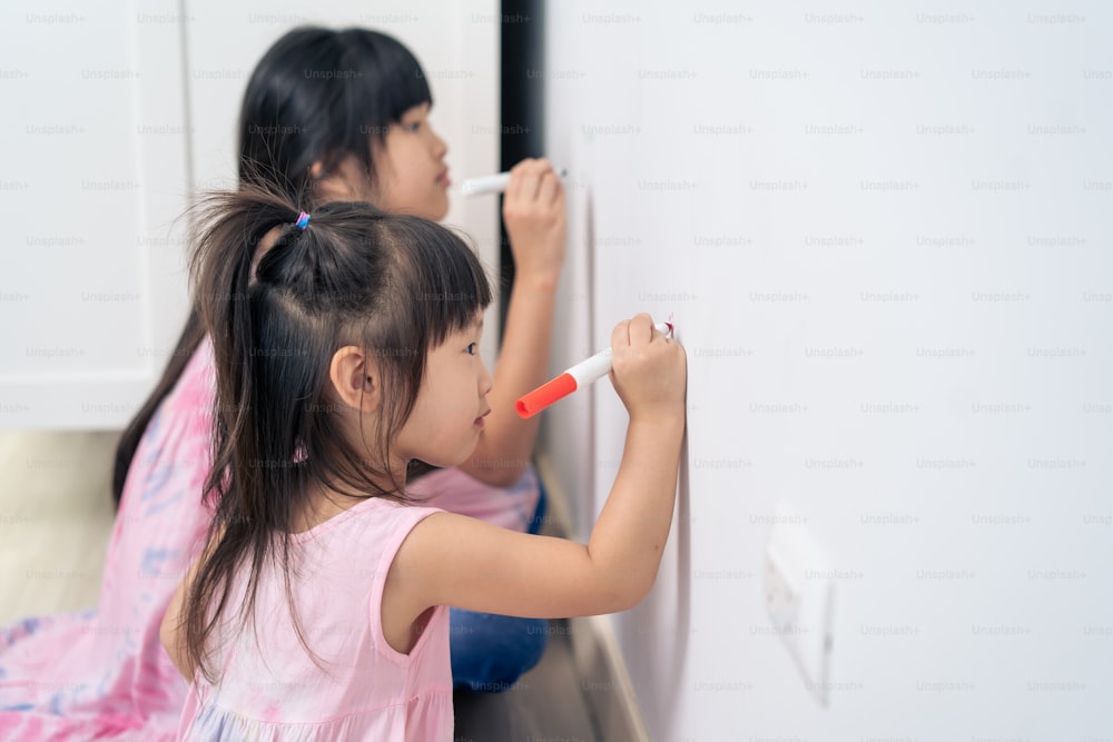 Asian young sibling kid girl enjoy paint on white wall in living room. Little adorable children having fun drawing and coloring art picture with hapiness enjoy creativity activity on holiday at home.