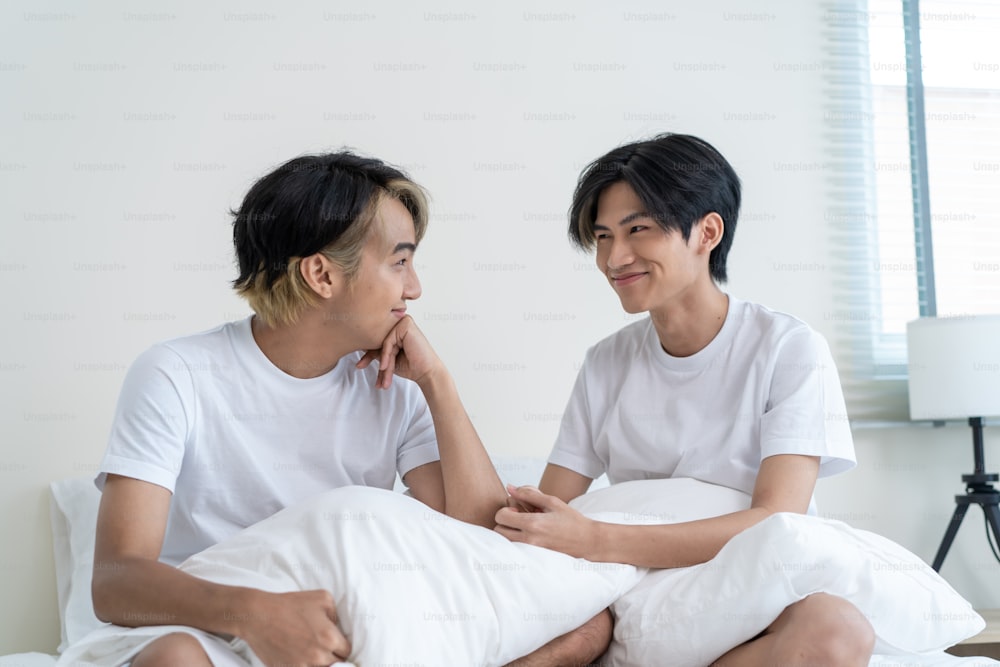 Portrait of Asian handsome gay couple sit on bed in bedroom at home. Attractive romantic male lgbt couple feeling happy to spend morning free time together and smile, looking at each other in house.