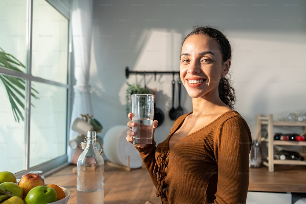 Joven hermosa mujer latina sosteniendo agua limpia en un vaso en la cocina. Atractiva bebida activa de la chica sedienta o tome un sorbo de mineral natural en taza para el cuidado de la salud y el bienestar en la cocina de la casa.