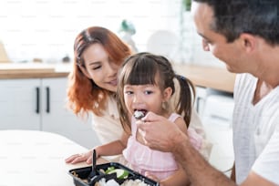 Asian attractive parents feeding food to cute baby daughter in kitchen. Happy family, loving couple cook and serve tasty healthy foods to little cute kid girl while sitting on chair for lunch in house