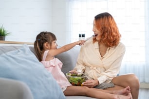Asian young happy kid daughter feed beautiful mom green salad in house. Beautiful woman mother enjoy eat vegetables healthy food to diet and lose weight for health care wellness with children in house