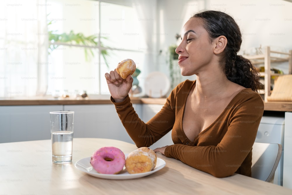 Latino attractive woman enjoy eat unhealthy foods in kitchen at home. Young beautiful girl sitting on dinner table, feeling hungry and happy to bite sweet chocolate donut and clean water in house.